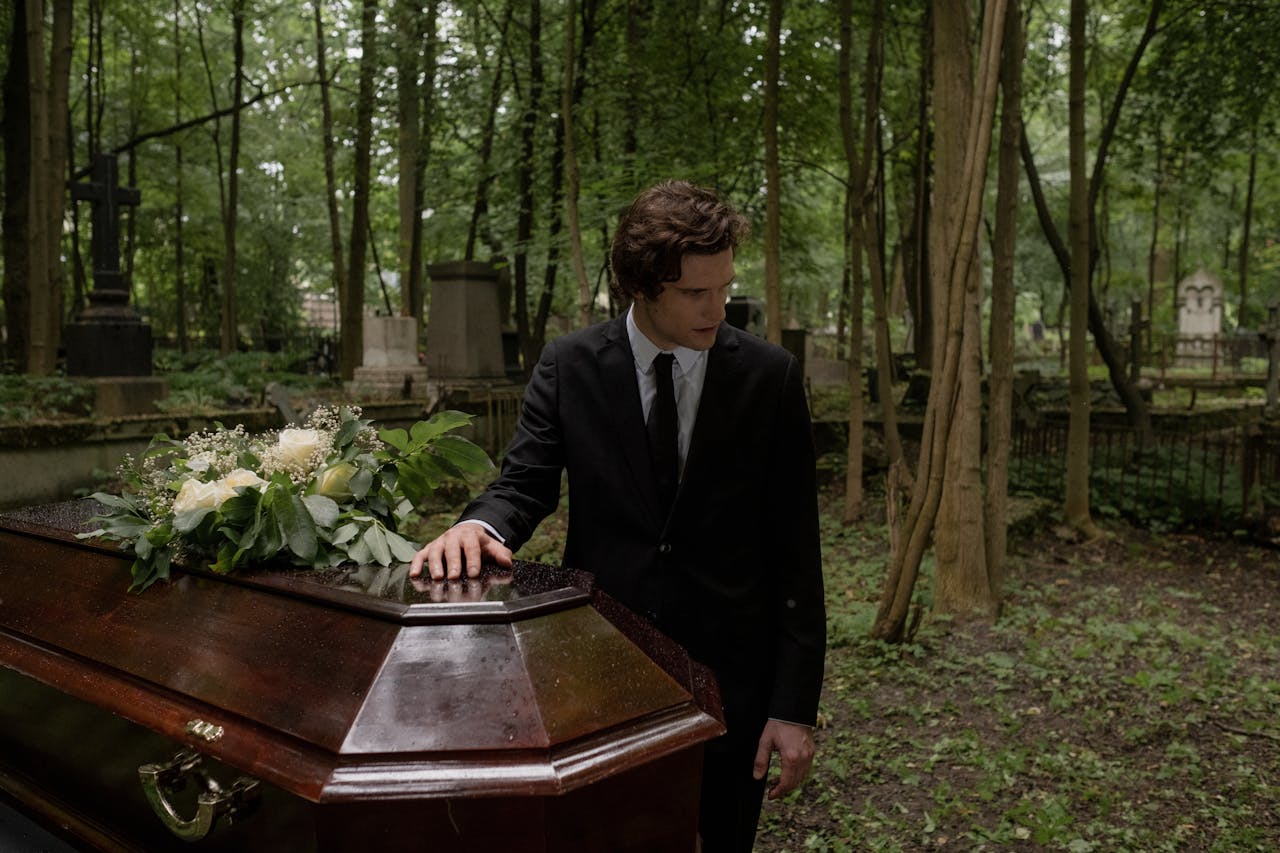 Home A man in a black suit mourns at a funeral in a lush green cemetery, leaning on the coffin.